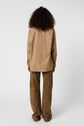 A person with curly hair stands facing away, wearing a loose chemise popeline camel and wide-legged brown pants against a plain white background.