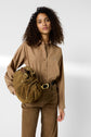 A woman with curly hair wears a chemise popeline camel and brown pants, holding a brown handbag. She stands against a plain light background, looking directly at the camera with a relaxed expression.