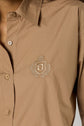 Close-up of a person wearing a chemise popeline camel, a tan button-up shirt with an embroidered crest featuring a shield, the letter J, and laurel branches on the chest. Only the shirt and part of the neck and shoulder are visible.