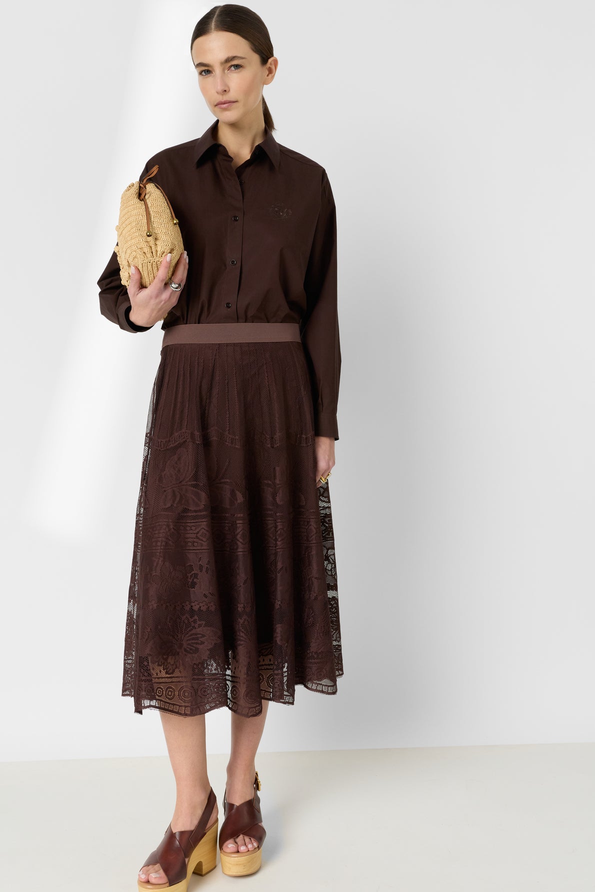 Woman in a brown button-up shirt, reminiscent of a chemise en popeline, and lace midi skirt holds a woven beige clutch. She wears brown platform sandals and stands against a plain white background. Her dark hair is pulled back.