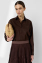 A woman with straight brown hair wears a dark brown chemise en popeline and matching skirt. She holds a woven beige clutch with gold accents against a plain light background.