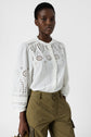 A person wearing a white, long-sleeve, eyelet blouse and olive green cargo pants stands against a plain, light background, looking confidently at the camera.