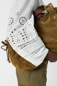 A person wearing a white eyelet blouse and olive pants holds a brown woven handbag with a dark brown strap and gold hardware.
