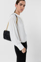 A person in a white blouse and black pants stands sideways, looking at the camera. They carry a black quilted handbag with a gold chain strap over their shoulder. The background is plain white.