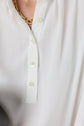 A close-up of a white blouse with four white buttons, partially unbuttoned at the collar. A gold chain necklace is visible around the neck above the blouse.