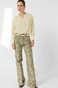 Woman standing against a white background, wearing a light yellow blouse en crêpe de soie with manches bouffantes, patterned flared pants, a beige belt, and brown shoes. She has earrings, her hair tied back, and a neutral expression.