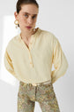 Woman in a pale yellow blouse jaune with buttons and floral patterned pants, crafted from crêpe soie and featuring elegant manches bouffantes, wearing large gold and pearl earrings, posing with hands behind her back against a plain light background.