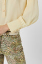 A person wearing a pale yellow crêpe soie long-sleeve blouse with subtle manches bouffantes and floral patterned pants with a beige belt, shown from mid-torso to upper thigh against a plain background.