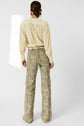 A person stands facing away, wearing a light yellow crêpe soie blouse with manches bouffantes and high-waisted, wide-leg floral pants in shades of green, yellow, and beige. Their hair is pulled back in a neat bun. The background is plain white.