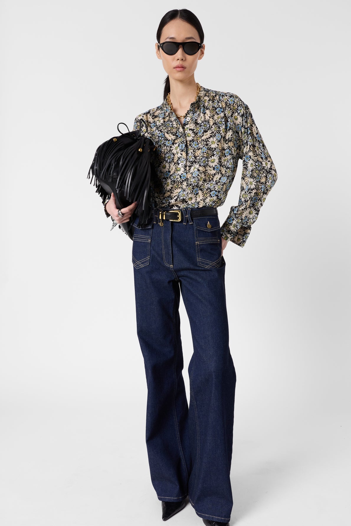 Woman wearing a motif floral blouse Clymène with a col tunisien, high-waisted dark blue wide-leg jeans, black pointed shoes, and black sunglasses, holding a black fringed handbag against a plain white background.