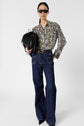 Woman wearing a motif floral blouse Clymène with a col tunisien, high-waisted dark blue wide-leg jeans, black pointed shoes, and black sunglasses, holding a black fringed handbag against a plain white background.