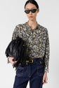 Woman wearing black sunglasses, a motif floral blouse Clymène with col tunisien, dark jeans, and gold jewelry holds a black fringed handbag. She stands against a plain light background, looking confidently at the camera.