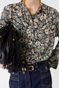 A person wearing a blouse Clymène with motif floral, blue jeans with white stitching, and a gold chain necklace holds a black fringed bag. Their hand rests on their hip against a plain background.