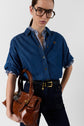 A person wearing a denim shirt over a striped top, dark jeans with a black belt, large glasses, and holding a brown leather handbag, stands against a plain background.