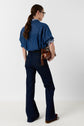 Woman with long brown hair in a ponytail, wearing glasses, blue denim shirt, dark blue jeans, and brown shoes, standing and holding a brown handbag, facing away from the camera against a plain background.