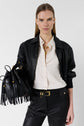 A woman wearing a black leather jacket, cream blouse, black pants, and gold accessories holds a black fringed handbag. She stands against a plain white background, looking directly at the camera.