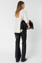 A woman with straight, light brown hair wears a white blouse and black leather pants, holding a black fringe handbag, standing against a plain white background, facing away from the camera.