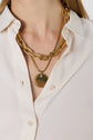 A person wearing a cream button-up shirt and two gold necklaces—one with large chain links and the other with a round pendant featuring a small gemstone.