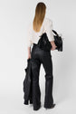 A person with straight, long hair wears a white top and black leather pants, holding a black fringe handbag and a black jacket, standing against a plain white background.