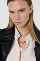 A woman with long blonde hair wears a black leather jacket over a cream shirt, layered gold necklaces, and looks directly at the camera with a neutral expression.