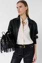 Woman in a black leather jacket and pants, white button-up shirt, and gold accessories holds a black fringed handbag. She has straight, light brown hair and looks to the side against a plain background.