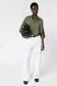 A person wearing a loose olive green shirt, white wide-leg pants, black belt, and black shoes holds a black handbag and wears chunky gold jewelry, standing against a plain white background.