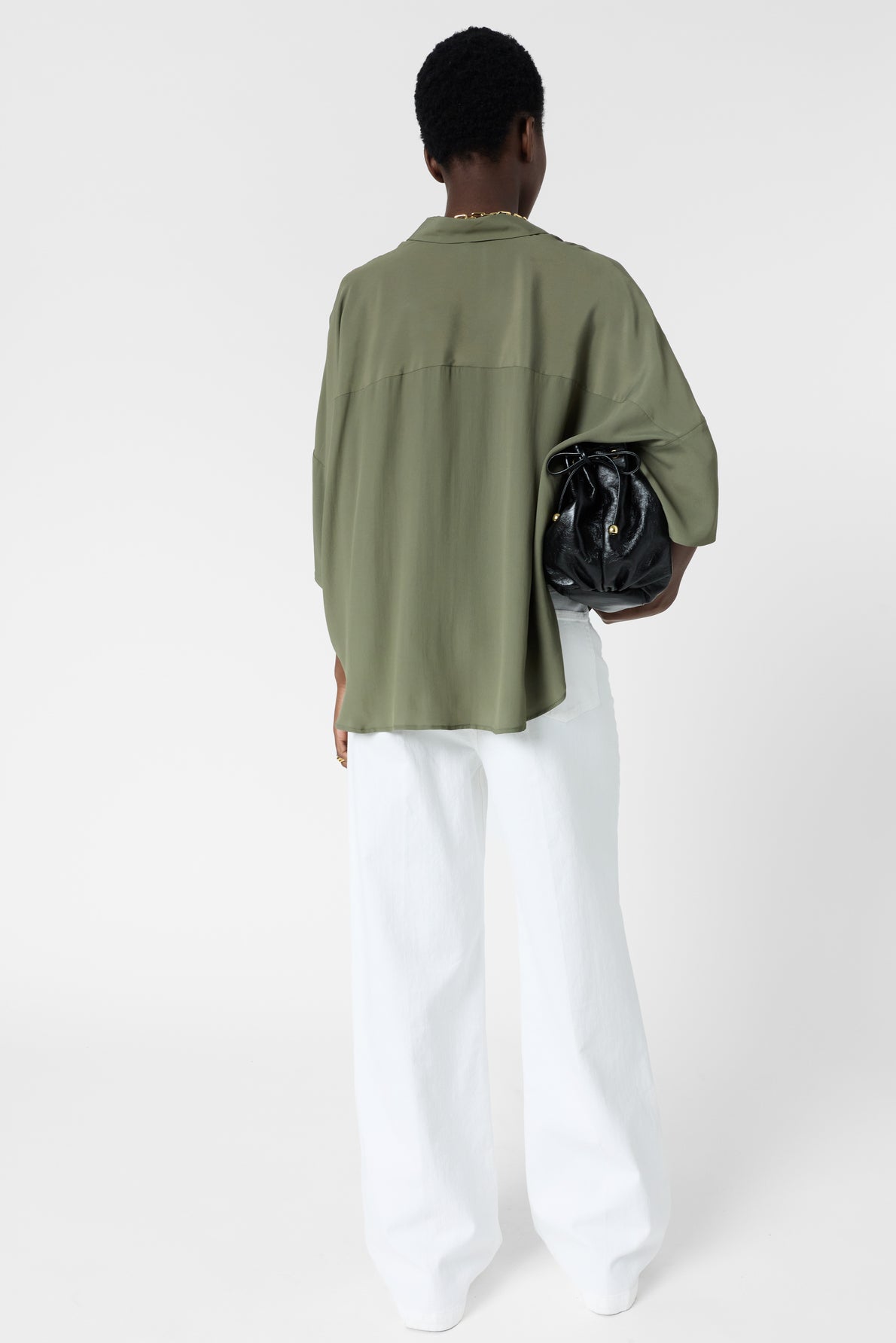 A person stands facing away, wearing a loose olive green top, white wide-leg pants, and holding a black handbag under one arm, against a plain white background.