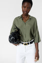 A woman in an olive green blouse and white pants holds a black handbag. She wears a chunky gold necklace and a black belt with a gold buckle. The background is plain white.