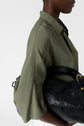 A person in an olive green shirt holds a black leather bag under their arm. The background is plain white. The photo is cropped to show only the lower face, torso, and bag.