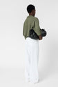 Person standing with back to camera, wearing an olive green shirt and white pants, holding a black handbag under one arm against a plain white background.