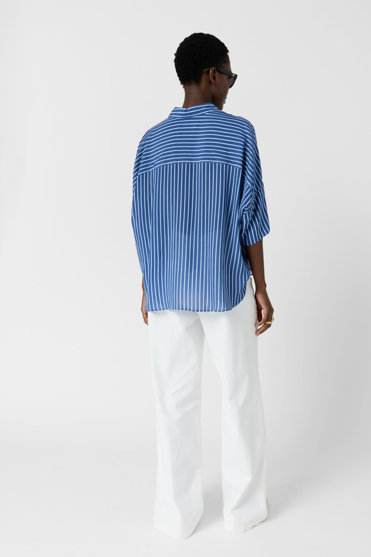 Person standing facing away, wearing a loose blue and white vertical striped shirt with wide sleeves, white pants, and dark sunglasses. The background is plain white.