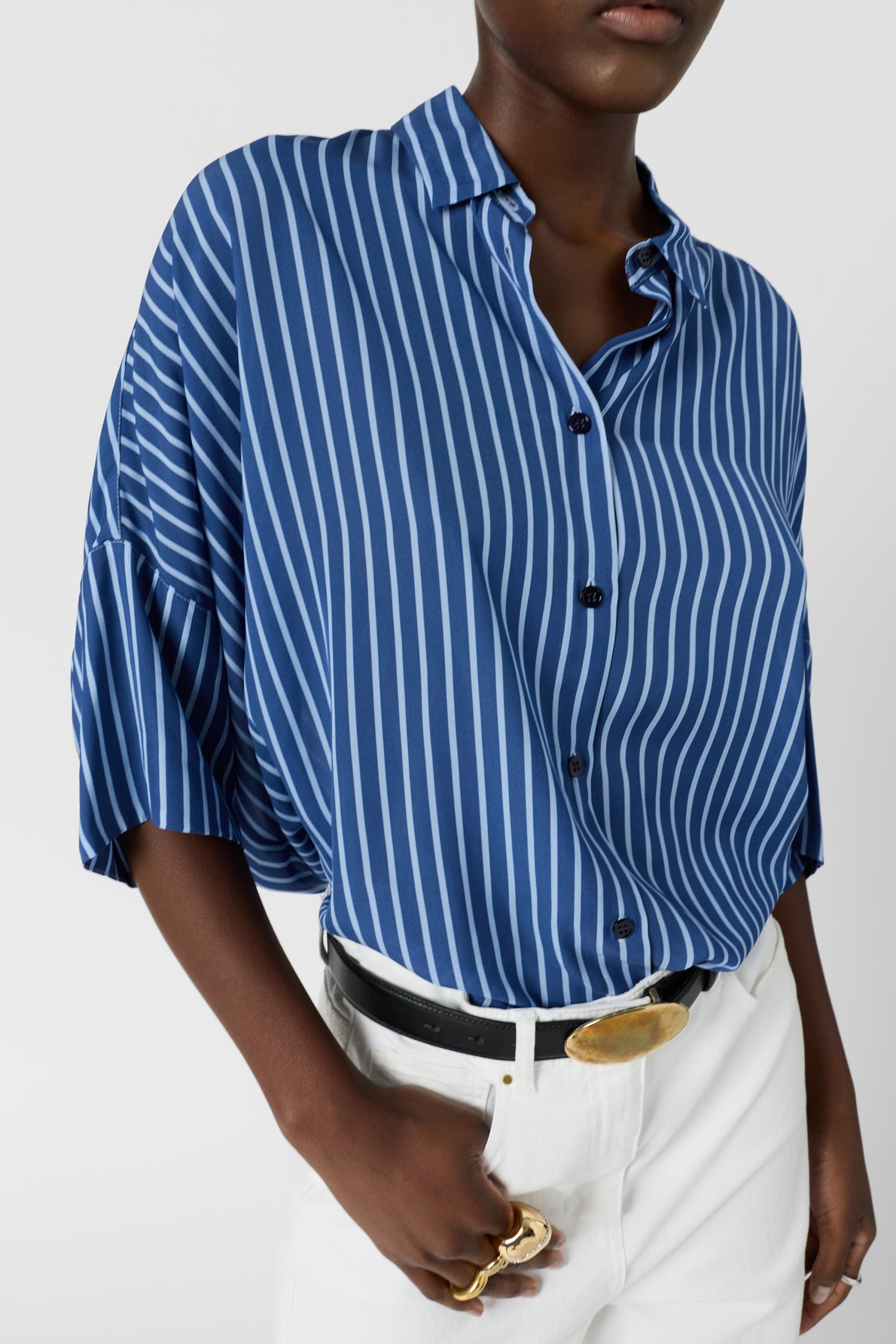 A person wearing a blue and white vertical striped button-up shirt tucked into white pants with a black belt and gold buckle, accessorized with gold rings.
