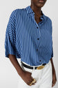 A person wearing a blue and white vertical striped button-up shirt tucked into white pants with a black belt and gold buckle, accessorized with gold rings.
