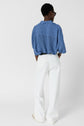 Person standing with back to camera, wearing a blue and white vertical striped shirt, white wide-leg pants, white shoes, and black sunglasses, against a plain white background.