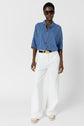 A person wearing a blue and white striped shirt, white wide-leg pants, white shoes, a black belt with a gold buckle, sunglasses, and gold jewelry stands against a plain white background.
