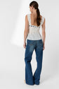 A woman with long brown hair in a ponytail wears an elegant white top dentelle sleeveless lace top and blue wide-leg jeans. She is standing and facing away from the camera against a plain white background.