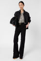 Woman stands against a white background wearing a black leather jacket, patterned blouse, black high-waisted pants, and tan shoes, holding a large black fringed bag under her arm.