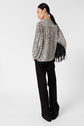 A woman with long black hair stands facing away, wearing a patterned blouse, black pants, and holding a black fringed handbag, against a plain white background.