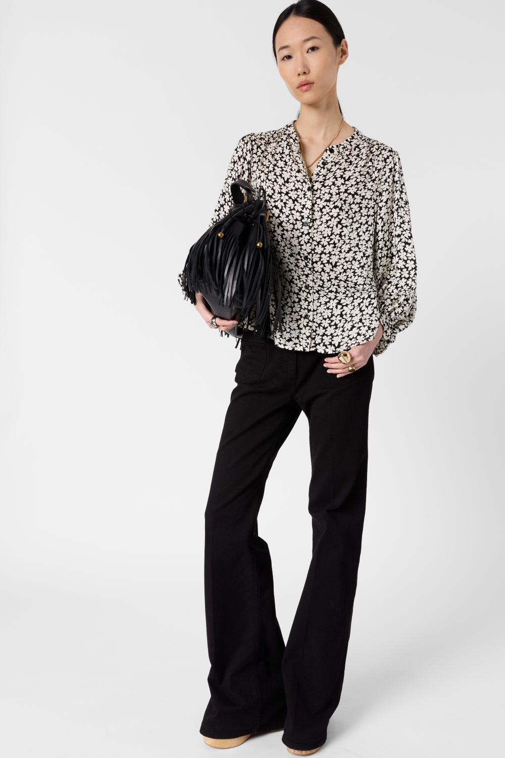 Woman standing against a plain background, wearing a black-and-white floral blouse, black wide-leg pants, and holding a black fringed handbag. She has straight hair pulled back and is looking at the camera.