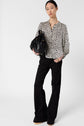 Woman standing against a plain background, wearing a black-and-white floral blouse, black wide-leg pants, and holding a black fringed handbag. She has straight hair pulled back and is looking at the camera.