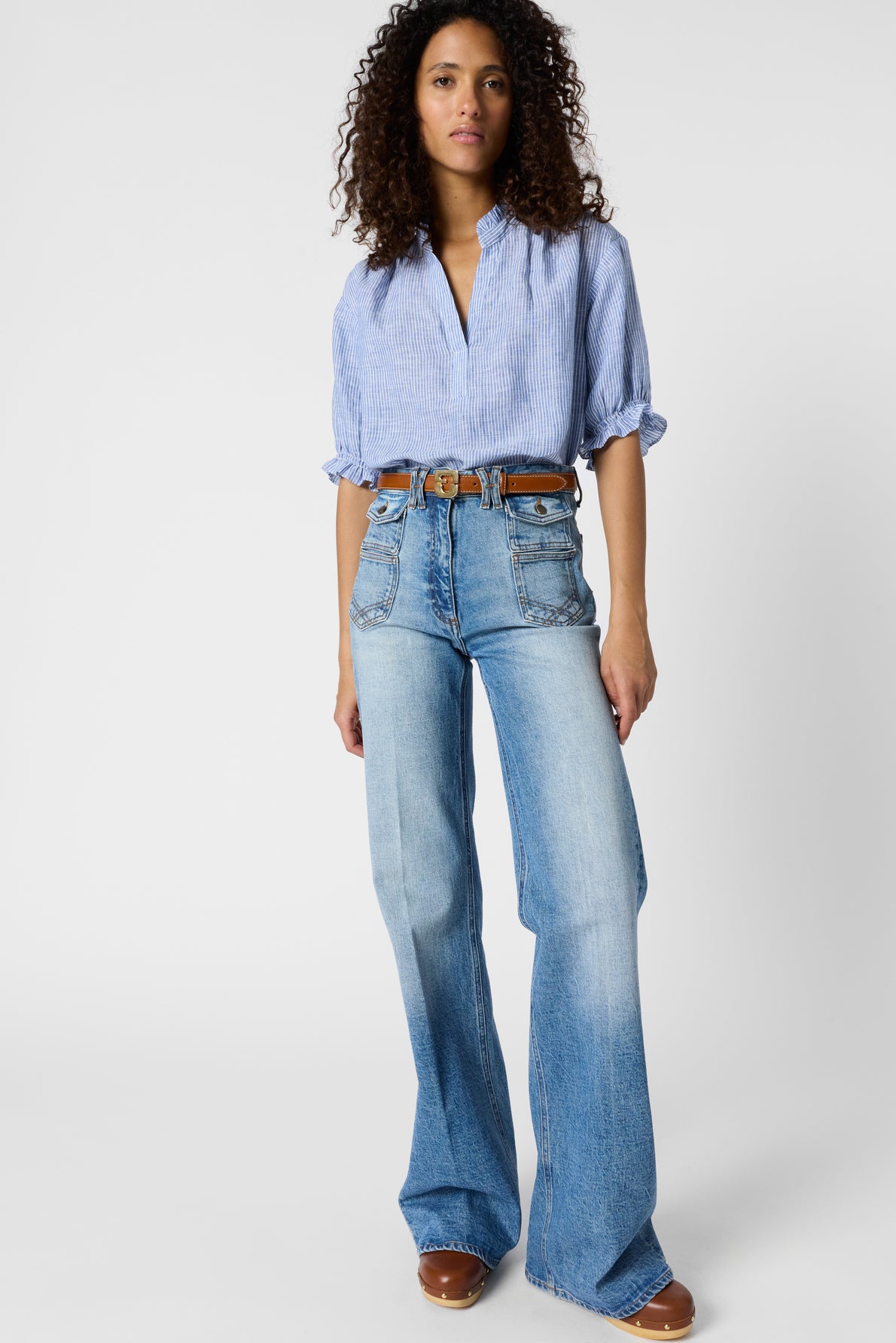A person stands facing forward, wearing a light blue, short-sleeve blouse femme buttoned up and tucked into high-waisted, light blue flared jeans. The look is completed with a brown belt and brown shoes against a plain background.