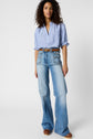A person stands facing forward, wearing a light blue, short-sleeve blouse femme buttoned up and tucked into high-waisted, light blue flared jeans. The look is completed with a brown belt and brown shoes against a plain background.