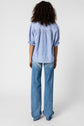 Person standing with back to the camera, wearing a light blue, short-sleeve, ruffled blouse à rayures and loose-fitting blue jeans that cover their shoes, against a plain white background.