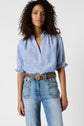 A person with curly hair wears a blue and white blouse à rayures tucked into high-waisted blue jeans with front pockets and a brown belt, standing against a plain white background.