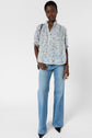 A person stands against a white background wearing a blouse fluide à col montant with a light blue motif floral and puff sleeves, paired with light blue flared jeans, pointed black shoes, and carrying a black shoulder bag.