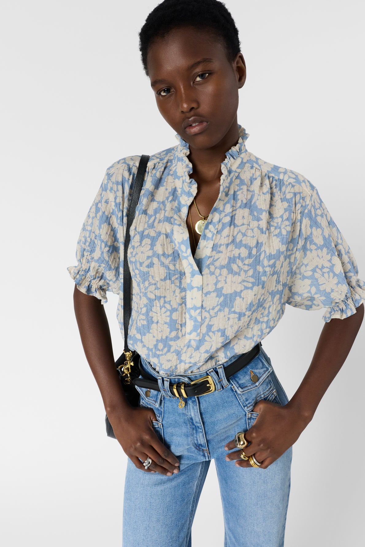 A person wearing a light blue blouse fluide à motif floral et col montant with high-waisted jeans stands with hands on hips, accessorized with rings, necklaces, and a black shoulder bag, against a plain white background.