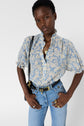 A person wearing a light blue blouse fluide à motif floral et col montant with high-waisted jeans stands with hands on hips, accessorized with rings, necklaces, and a black shoulder bag, against a plain white background.