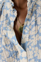 A person wearing a blue and beige motif floral blouse fluide with a col montant ruffled collar, showcasing two gold necklaces, one with a round pendant and a small gemstone detail.