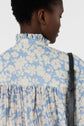A person with short curly hair is shown from behind, wearing a light blue and white blouse fluide with a motif floral design, featuring a ruffled col montant and a black shoulder strap.