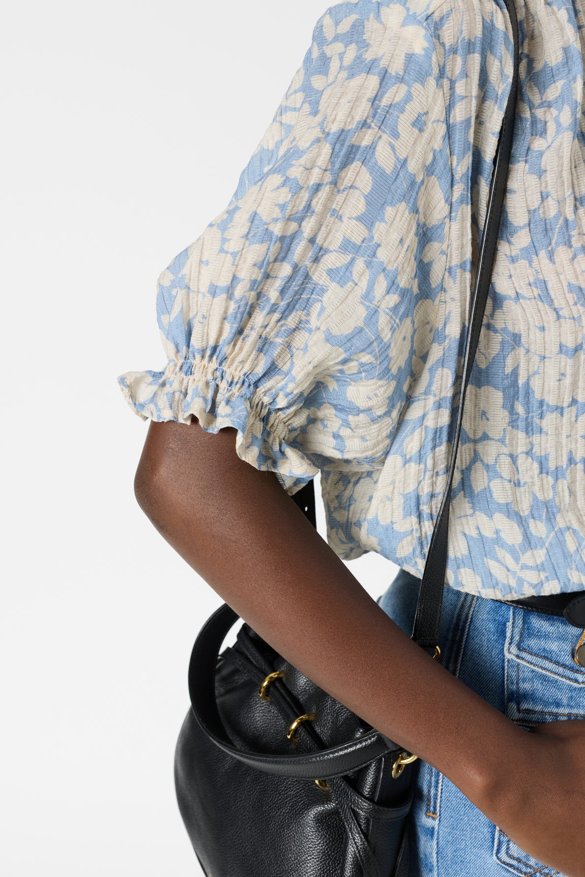 A person wearing a blue and white blouse fluide with a delicate motif floral design and blue jeans holds a black leather bag over their shoulder. The image focuses on their arm, blouse sleeve, and the bag.